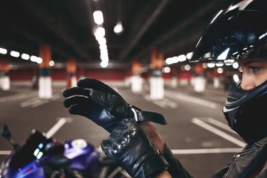Confident young female racer wearing stylish motorcycle helmet putting on leather gloves, posing isolated in underground parking lot with her blue motorbike. Selective focus on woman's hands © Freepik